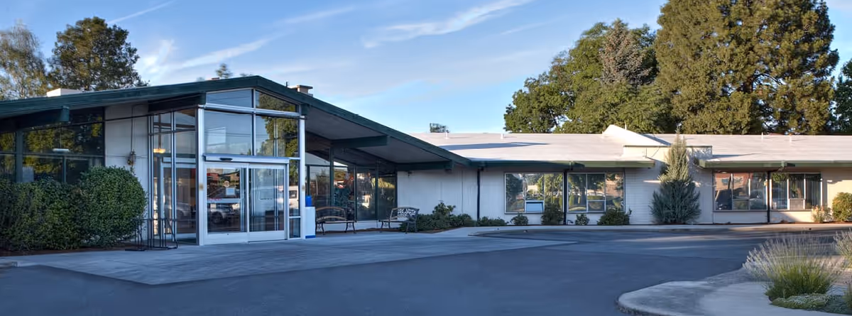 Exterior view of a single-story building with large windows and a glass entrance door, surrounded by bushes and trees under a blue sky with light clouds.