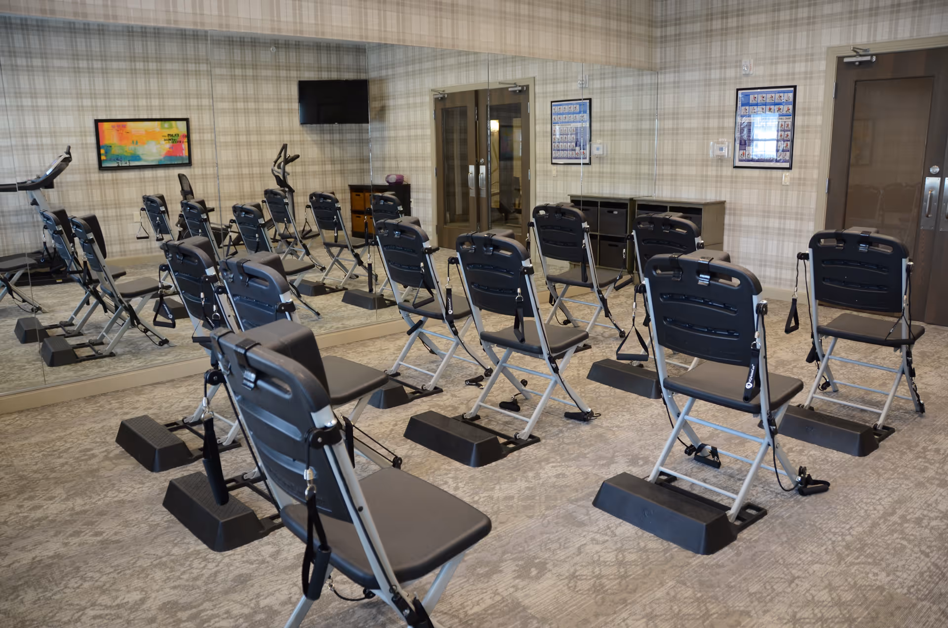 Exercise studio with rows of folding exercise chairs equipped with resistance bands facing a mirrored wall and a TV.