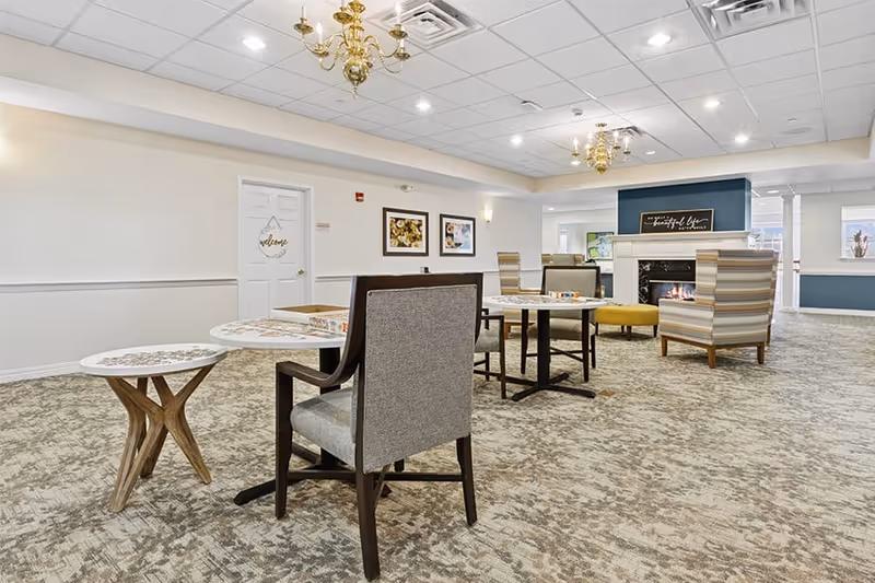 A spacious and well-lit common area with carpeted flooring, several tables and chairs arranged for seating, a fireplace with a decorative sign above it, and framed artwork on the walls. The ceiling has recessed lighting and chandeliers, and there is a door with a welcome sign hanging on it.