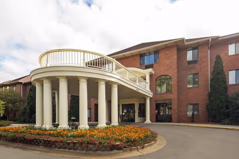 Front entrance of a red-brick senior living building featuring a white columned circular portico and landscaped flower beds.