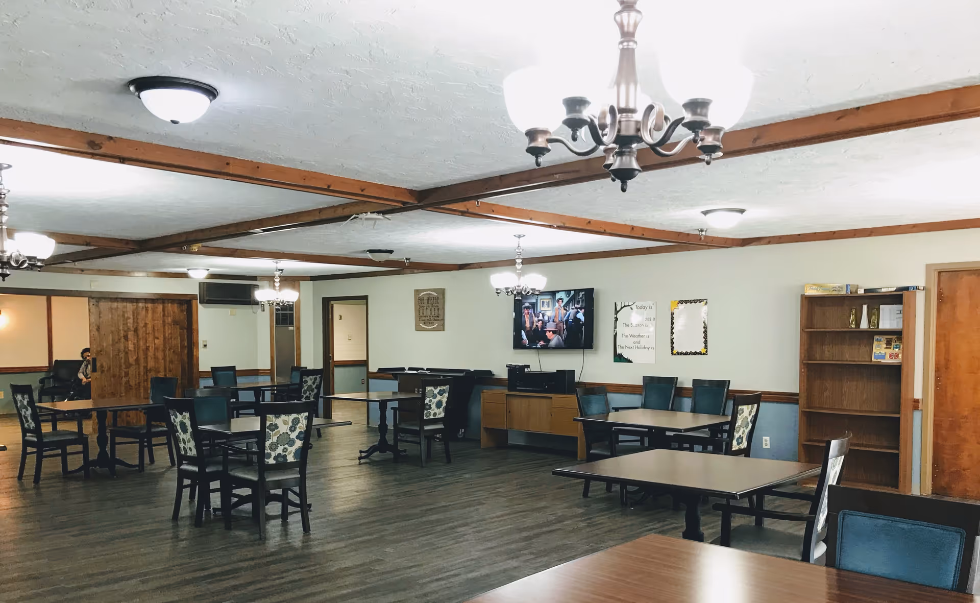 Spacious communal dining/activity room with multiple tables and chairs, chandeliers, a wall-mounted TV, and shelving.