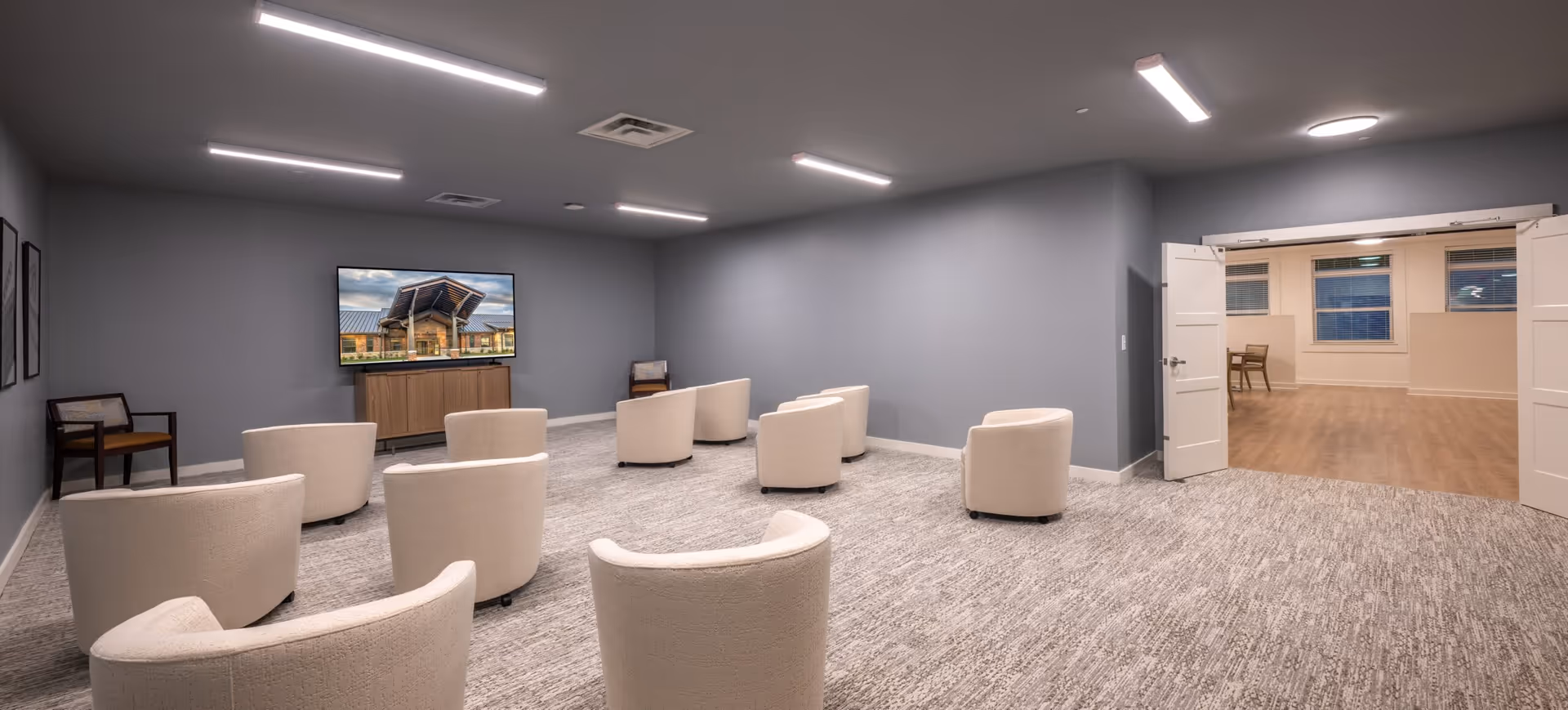 A modern senior living facility common room with multiple beige armchairs arranged in rows facing a large flat-screen TV mounted on a gray wall. The room has gray walls, carpeted flooring, and bright overhead lighting. Double doors open to an adjacent room with wooden flooring and a table with chairs near windows.