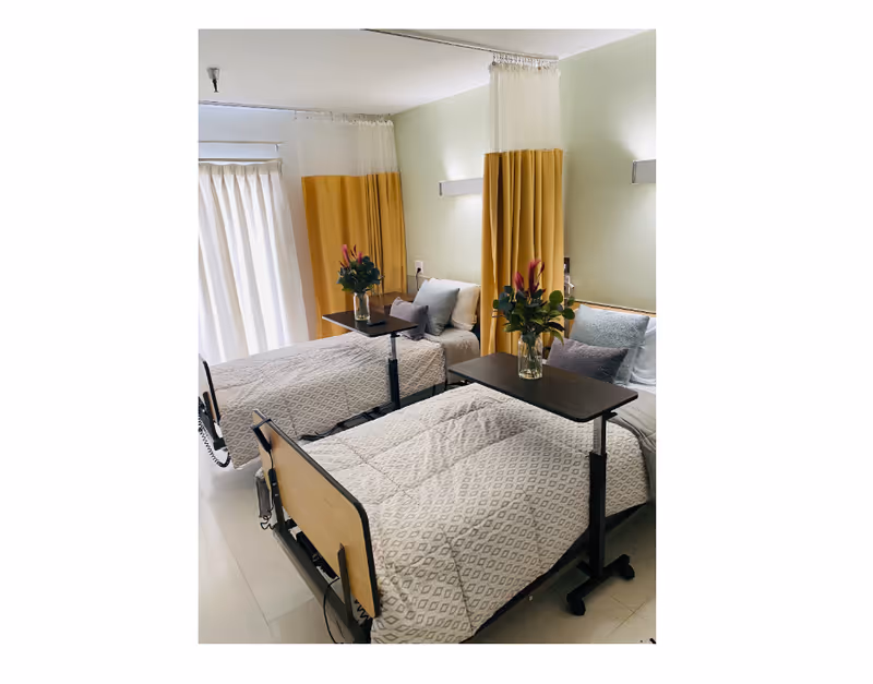 Two hospital-style beds with privacy curtains, tables, and vases of flowers in a bright room.