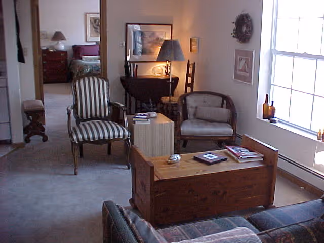 A cozy living room area with a wooden chest used as a coffee table, two armchairs with different upholstery, a small side table with a lamp, and framed artwork on the walls. A large window lets in natural light. In the background, a bedroom with a bed and nightstand is visible through an open doorway.