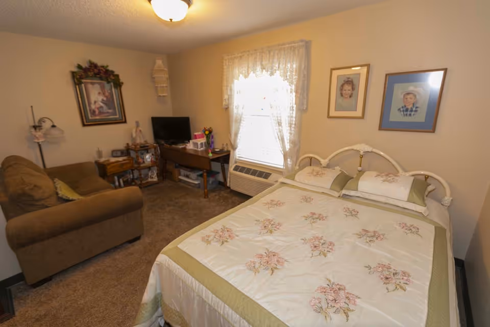 A cozy bedroom with a floral bedspread, a sofa, a small TV on a desk, and framed portraits on the walls.