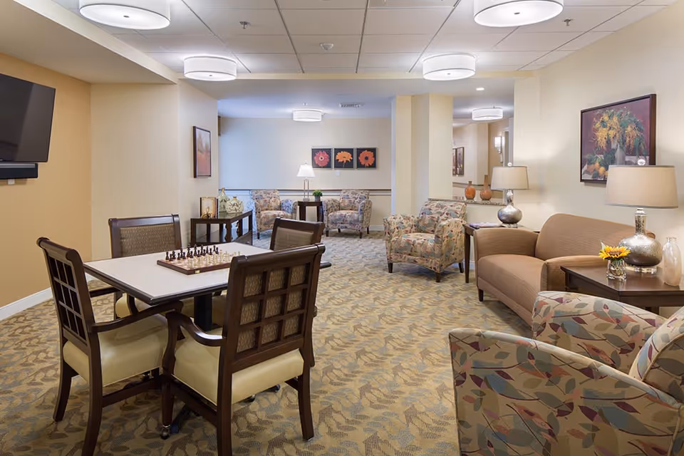 A cozy senior living common area with patterned armchairs, a brown sofa, side tables with lamps and flowers, a table with a chessboard, and framed floral artwork on the walls. The room has soft lighting from ceiling fixtures and a wall-mounted TV.