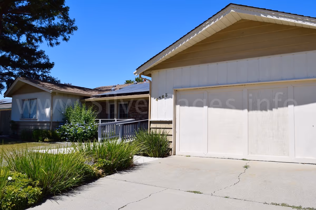 Exterior view of a single-story residential building with a beige garage door, a driveway, and a small garden with green plants and bushes. The house has a sloped roof and a ramp leading to the entrance. The sky is clear and blue.