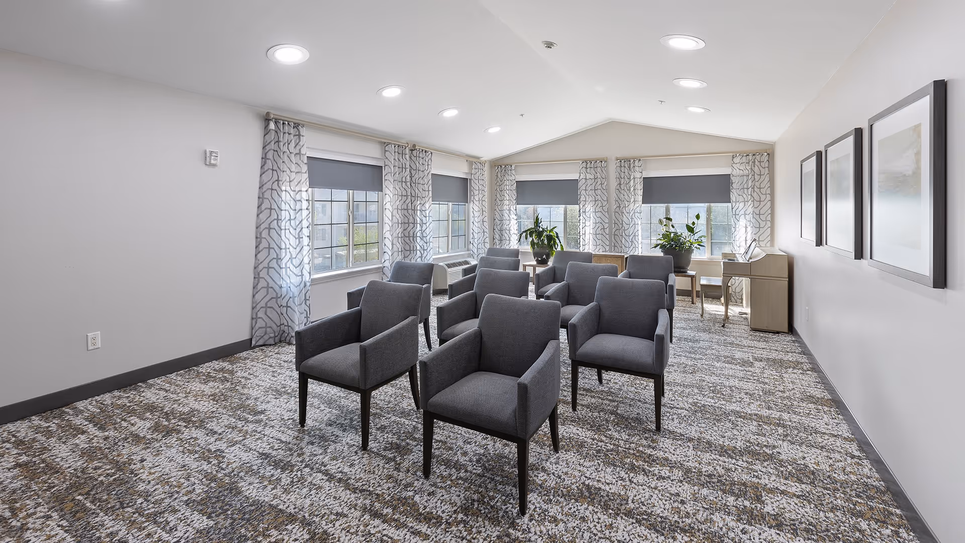 A bright room with multiple gray upholstered armchairs arranged in rows facing a wall with three framed pictures. The room has large windows with patterned curtains and gray roller shades, a patterned carpet, and a small desk with a chair and plants near the windows.