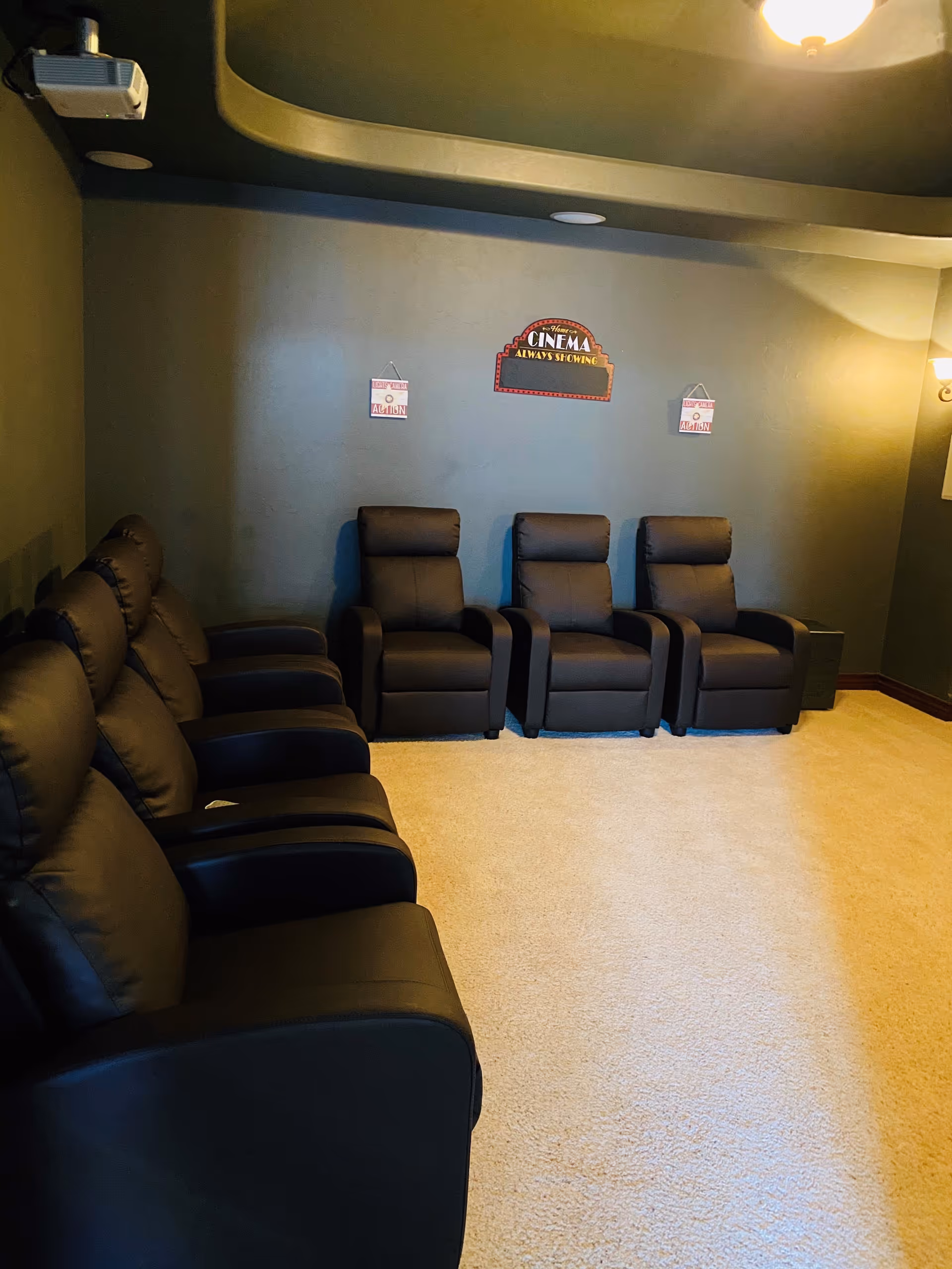 A small home theater room with six black leather recliner chairs arranged in two rows facing a blank wall with a projector mounted on the ceiling. The walls are painted dark green, and there are three decorative signs on the wall, including one that says 'Home Cinema Always Showing.' The room is carpeted in light beige.