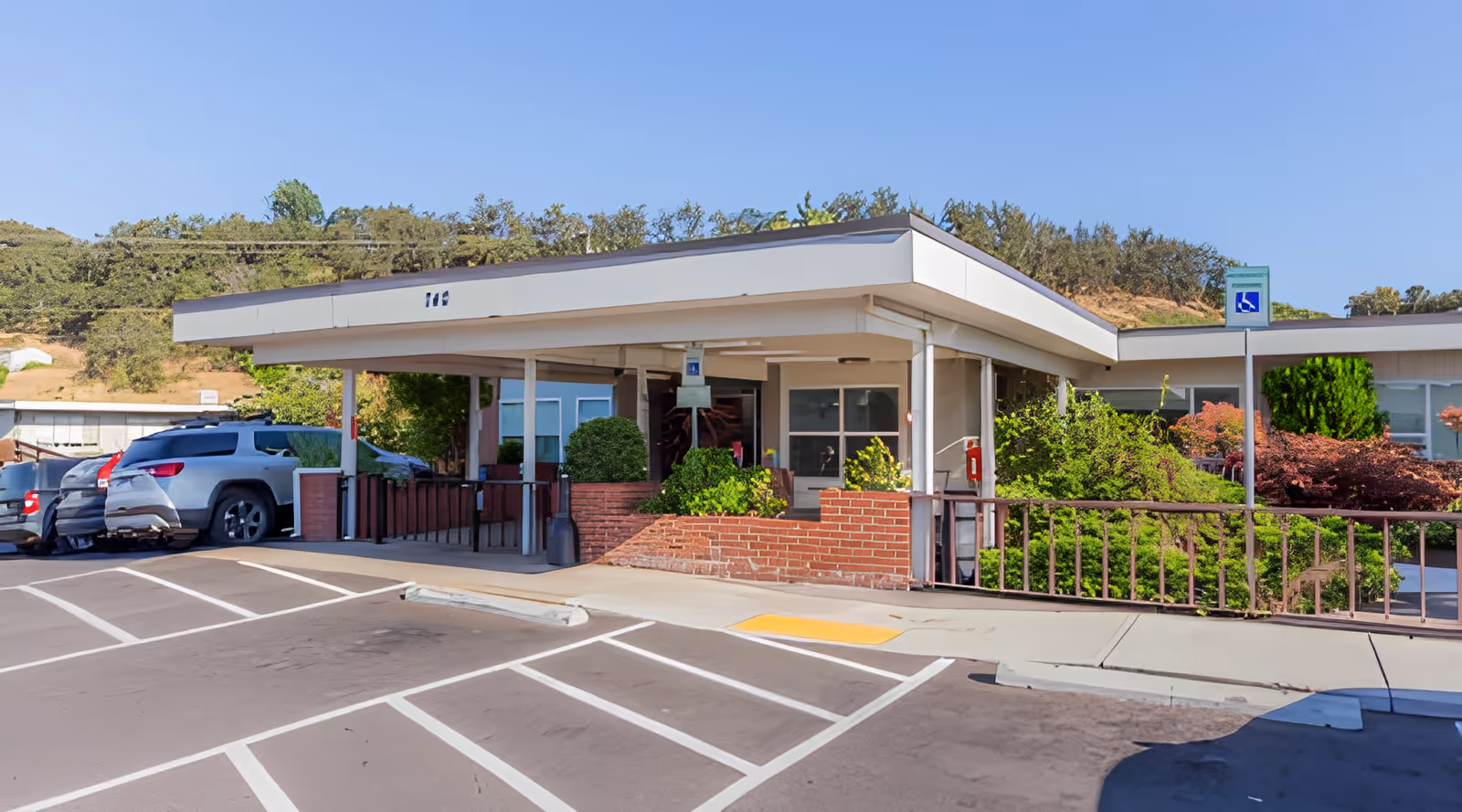 Entrance of Rose Haven Nursing Center showing a covered drop-off area with a ramp, surrounded by greenery and parked cars in designated parking spaces including a handicapped spot.