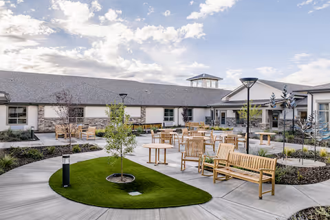 Outdoor courtyard area at Kiley Ranch Senior Living featuring wooden benches and tables arranged on a paved walkway with small landscaped green patches and young trees under a partly cloudy sky.
