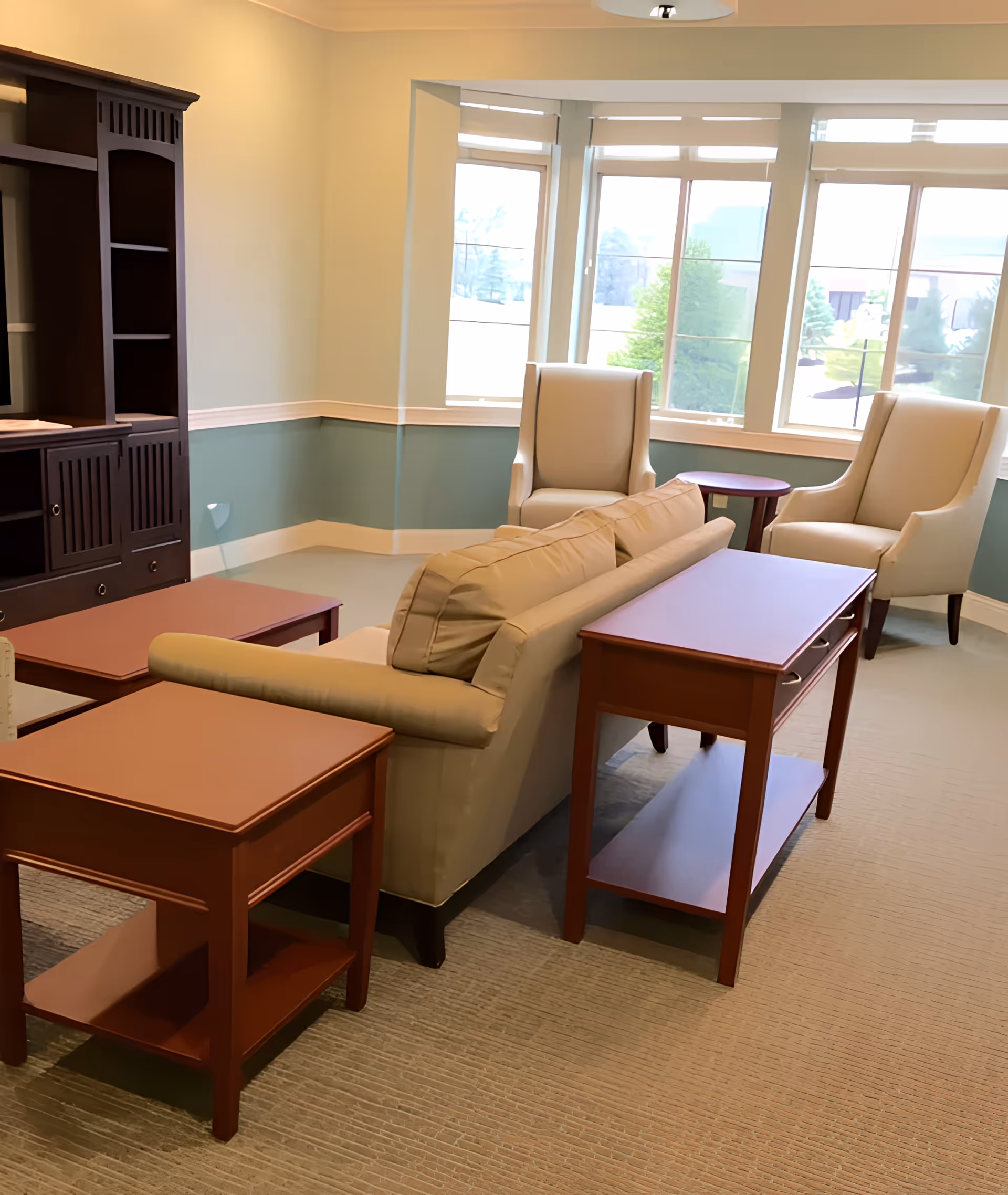 A cozy living room area with a beige sofa, two beige armchairs, and several wooden tables. Large windows allow natural light to fill the room, and a dark wooden entertainment center is visible on the left side.