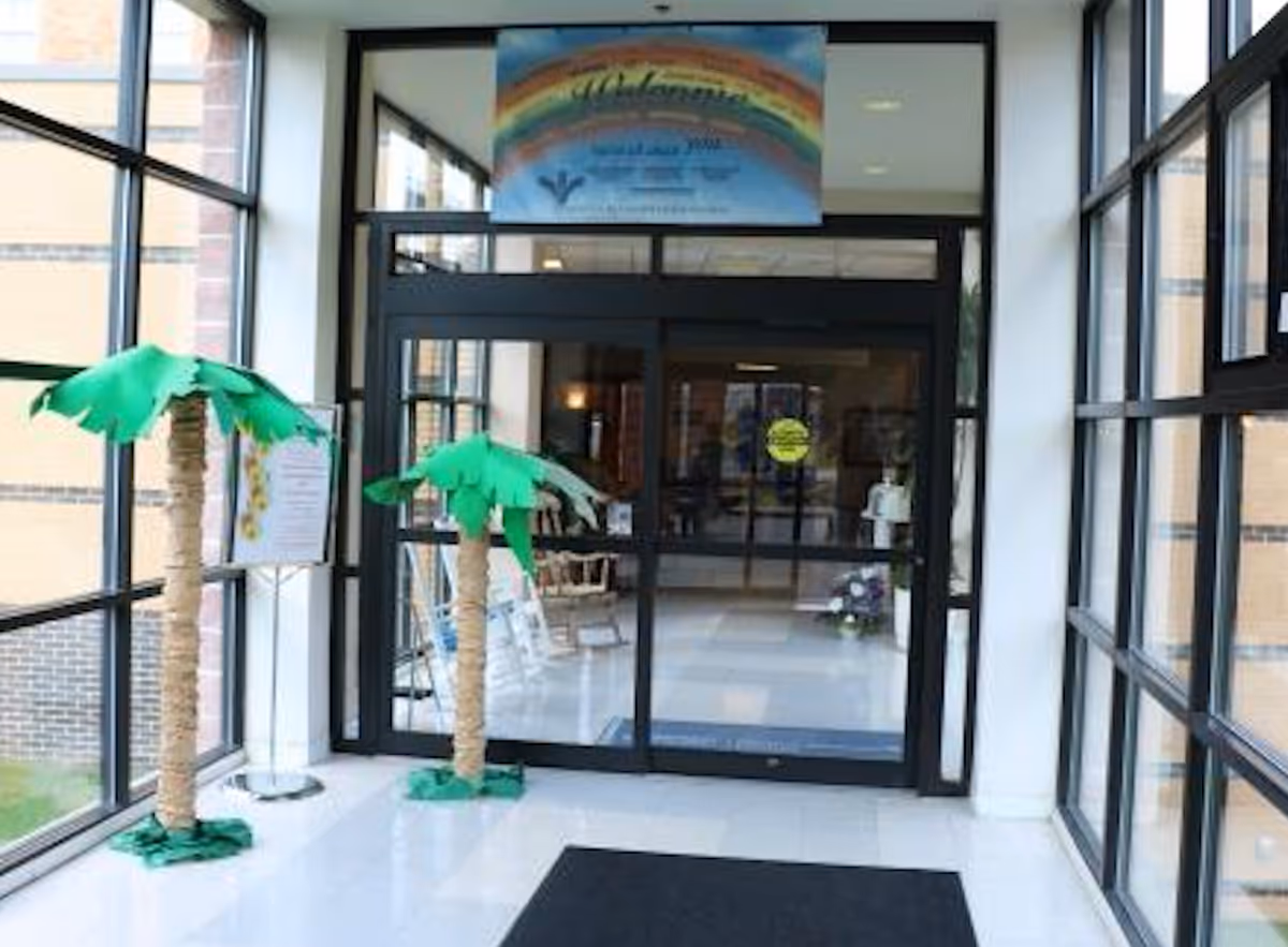 Entrance area of Mt Alverno Center with large glass windows on both sides, two decorative artificial palm trees, a black floor mat, and automatic sliding glass doors leading inside. Above the doors is a colorful welcome sign with a rainbow design.
