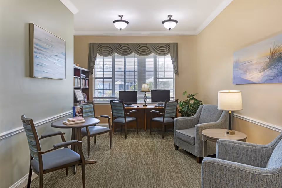 A cozy interior room with two gray armchairs and a small round table with a lamp between them on the right side. On the left side, there is a small round table with two chairs. At the back, a large window with blinds and a valance lets in natural light. Below the window, a desk with two computer monitors and two chairs is visible. The walls are painted in light beige and green tones, decorated with framed artwork and a bookshelf filled with books.
