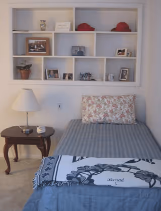 A small bedroom with a single bed covered in a blue striped bedspread and a floral pillow. A white and black patterned blanket is folded at the foot of the bed. Next to the bed is a wooden side table with a white lamp and a small clock. Above the bed is a white wall-mounted shelf with various framed photos, decorative items, and two red hats.