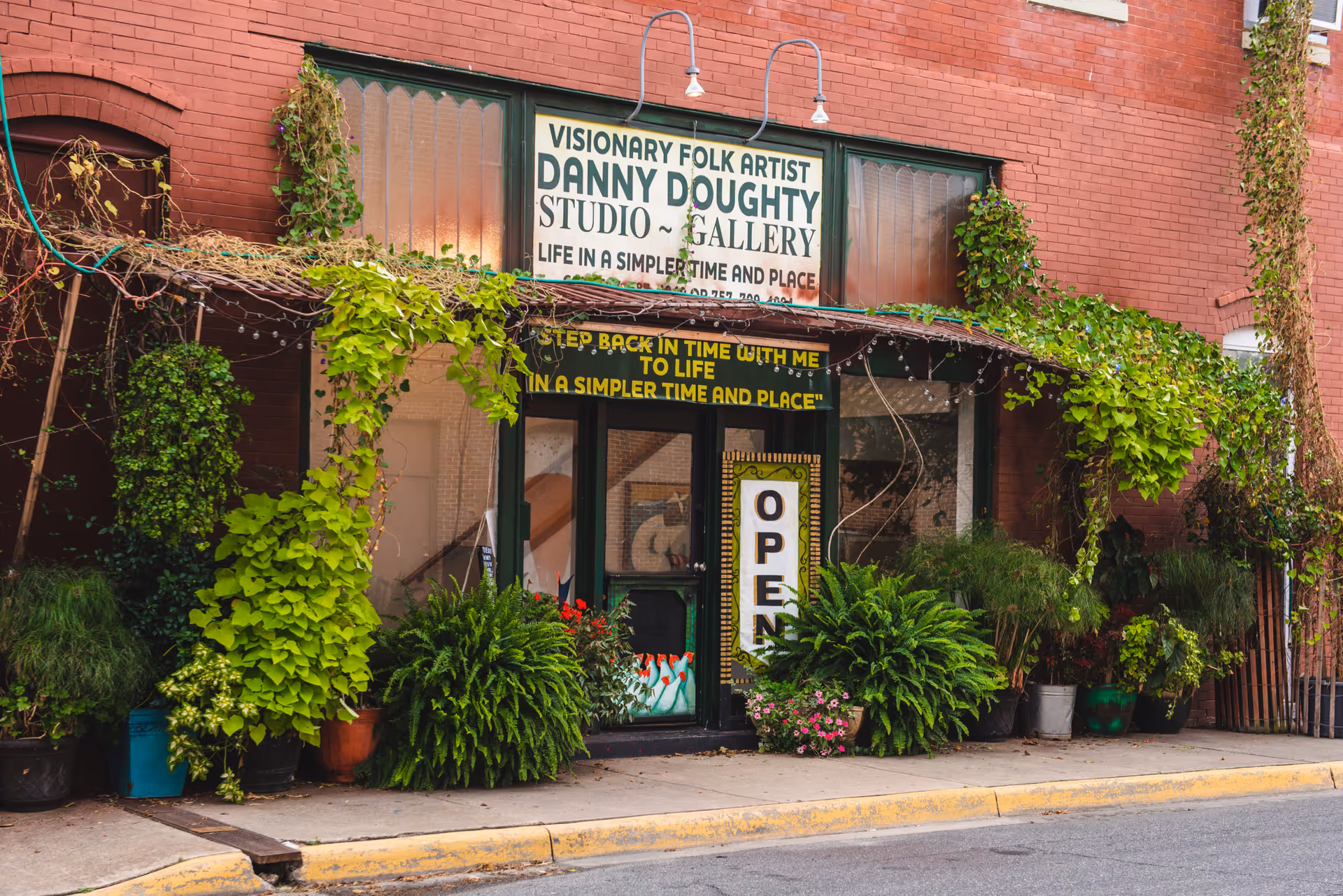 Exterior view of a brick building with a storefront covered in green vines and various potted plants. The storefront has a sign that reads 'Visionary Folk Artist Danny Doughty Studio ~ Gallery Life in a Simpler Time and Place' and a vertical 'OPEN' sign on the door.