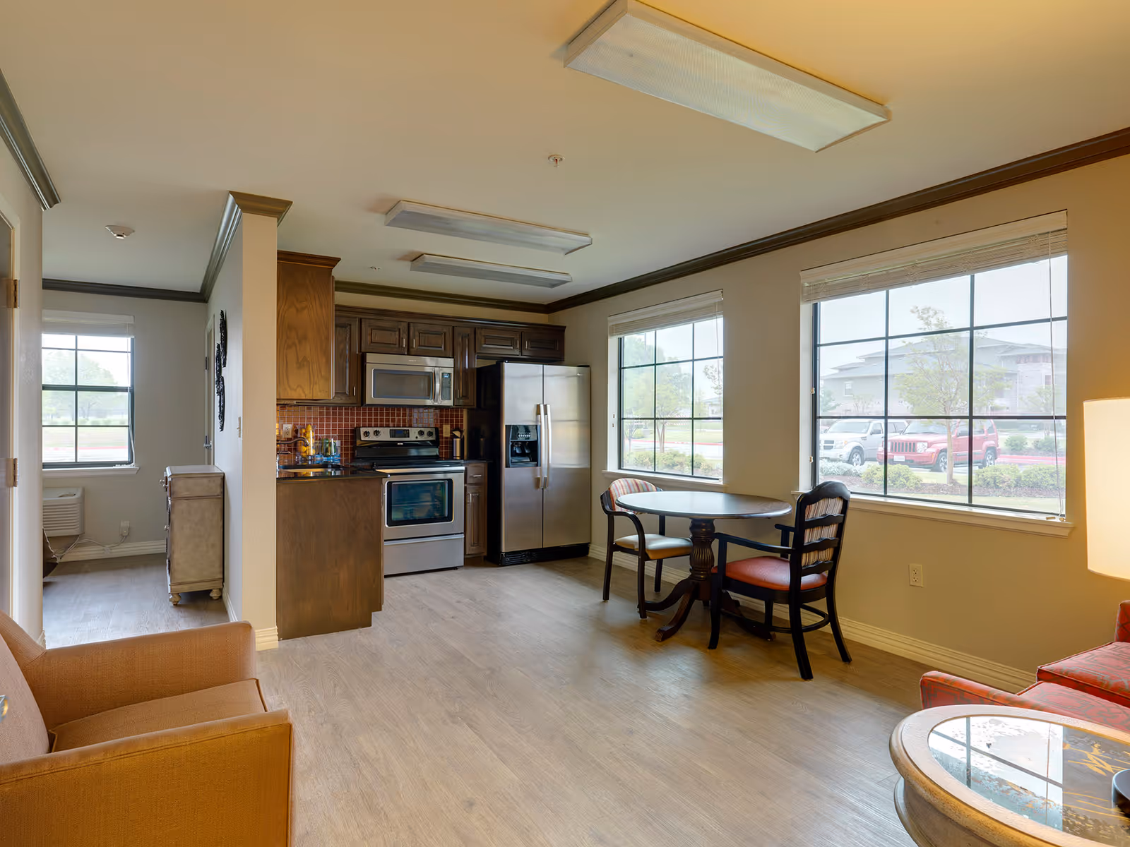 Interior view of a senior living facility kitchen and dining area with wooden cabinets, stainless steel appliances including a refrigerator and oven, a small round dining table with two chairs near large windows, and part of a living room with an armchair and a small table.