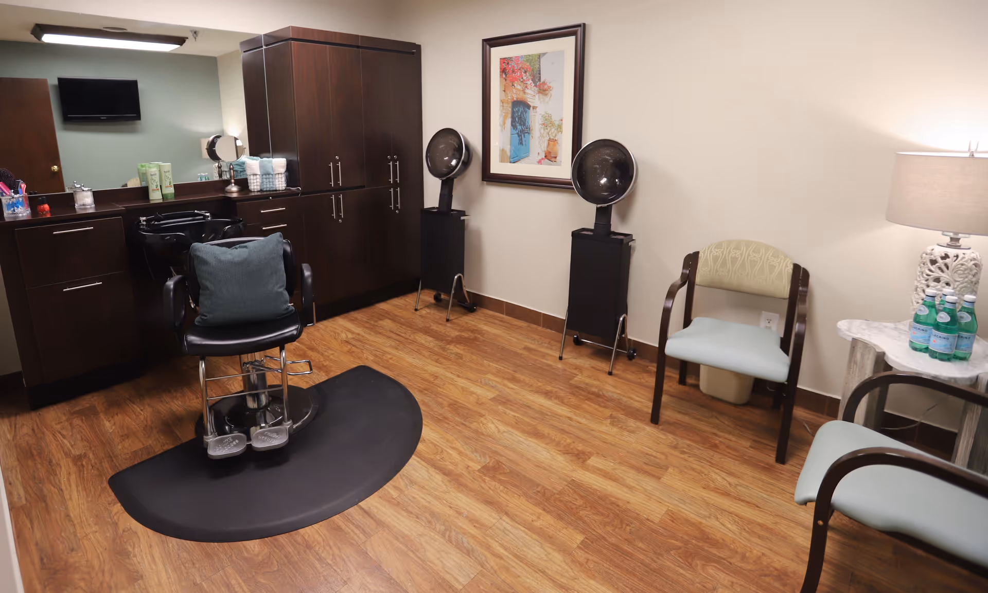 A small salon-style room with a styling chair and sink, dark cabinets, two hooded hair dryers, and guest chairs with a side table and lamp.