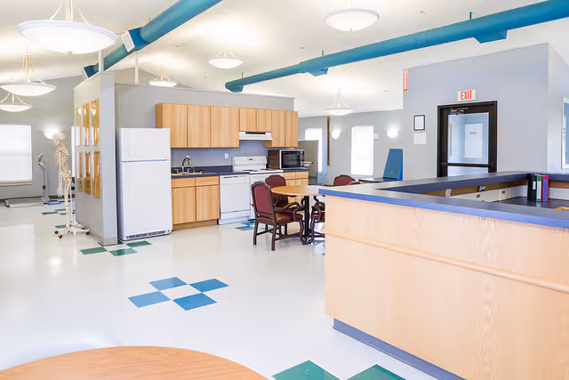 Bright communal room with a kitchenette, dining table, chairs, and a reception counter under blue exposed ducts.