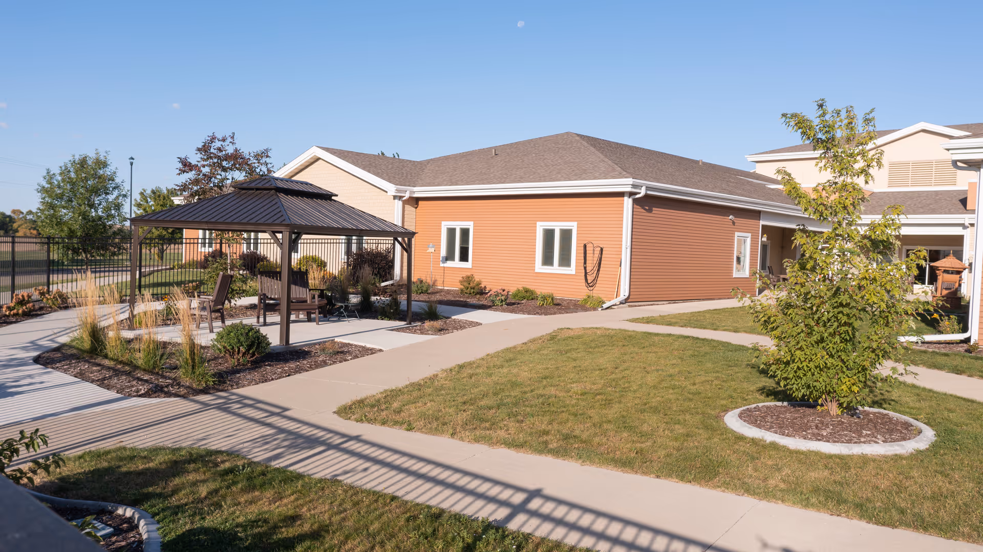 Outdoor area of North Dakota Veterans Home featuring a paved walkway, a small gazebo with seating, landscaped garden beds, and a building with orange siding under a clear blue sky.