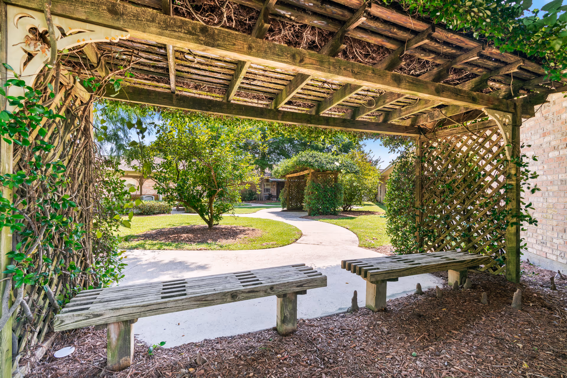 A shaded wooden pergola with two wooden benches underneath, surrounded by greenery including trees and bushes. A curved concrete pathway leads through a well-maintained garden area with more pergolas and a brick building visible in the background under a clear blue sky.