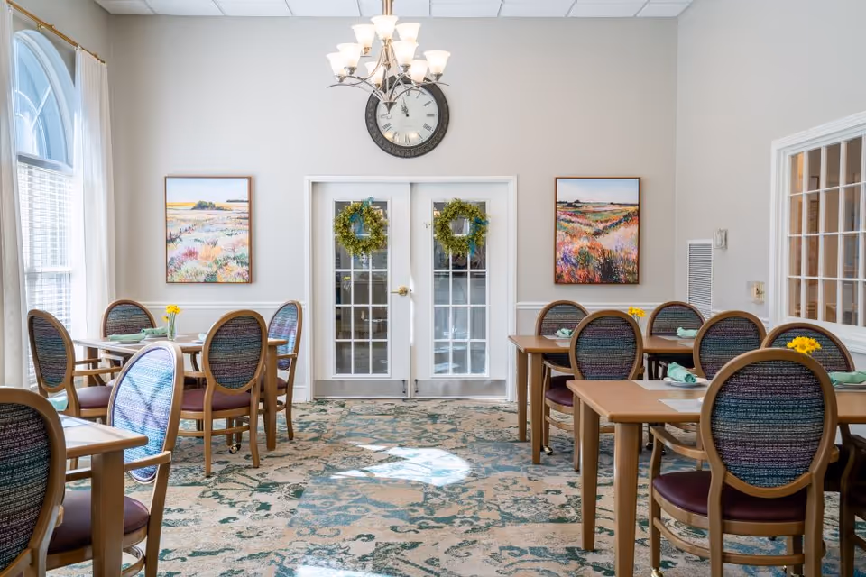 Bright dining room with multiple wooden tables and upholstered chairs, double glass doors adorned with wreaths, a wall clock, and colorful landscape paintings.