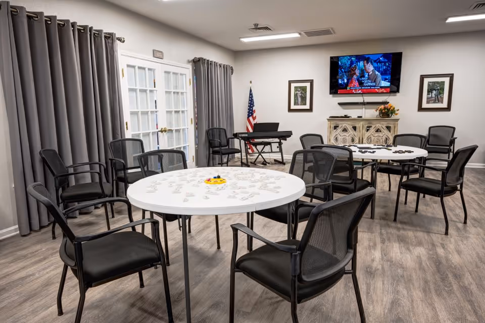 A well-lit activity room with two round tables surrounded by black mesh chairs. The tables have game pieces on them. The room features gray curtains covering windows, a keyboard on a stand, an American flag, framed pictures on the wall, and a flat-screen TV mounted above a decorative cabinet.
