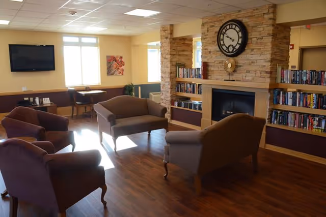 Cozy senior living common room with armchairs and loveseats arranged around a fireplace with built-in bookshelves and a wall clock.
