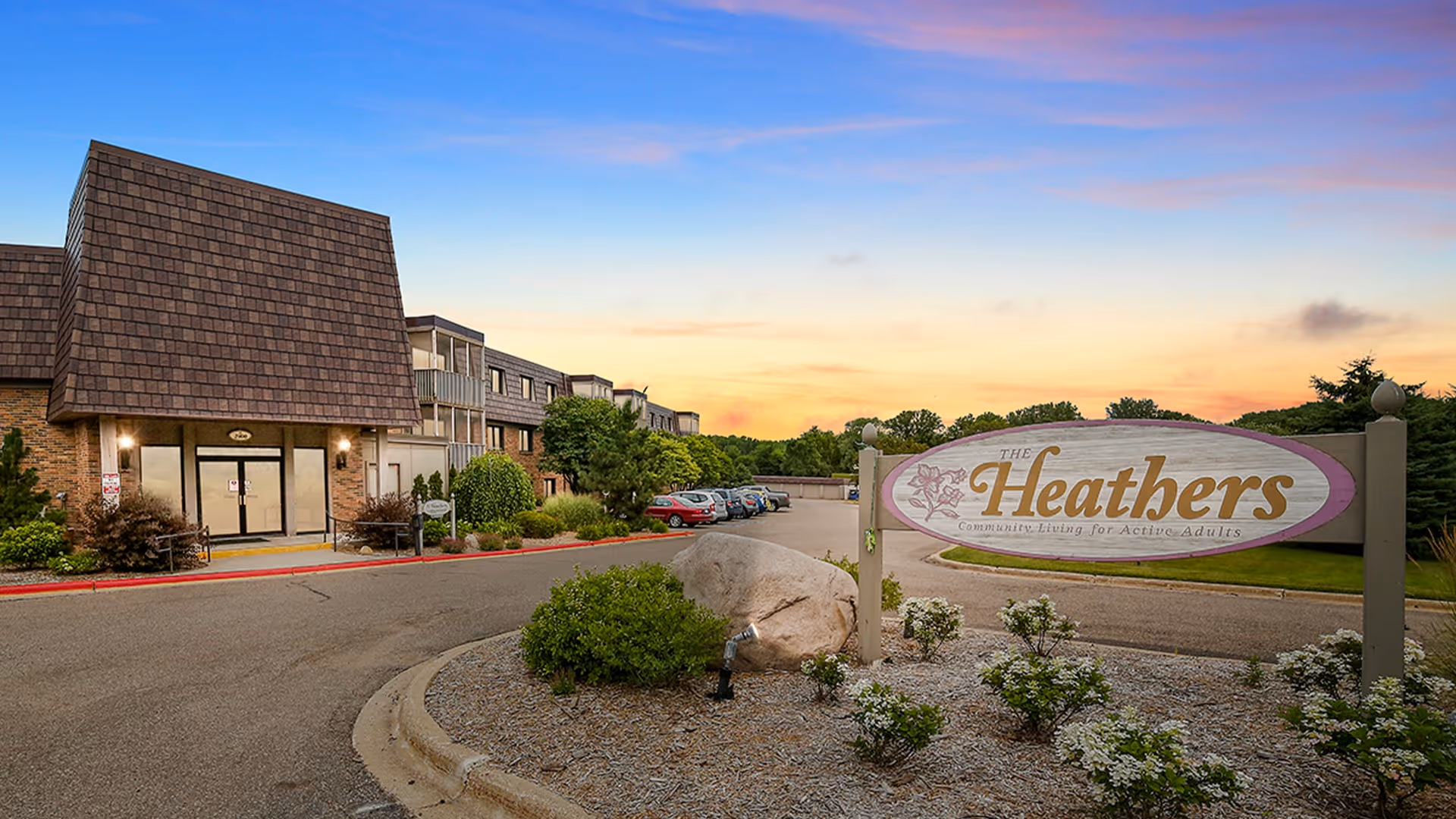 Front entrance and sign for The Heathers senior living community with a driveway and landscaped grounds at sunset.
