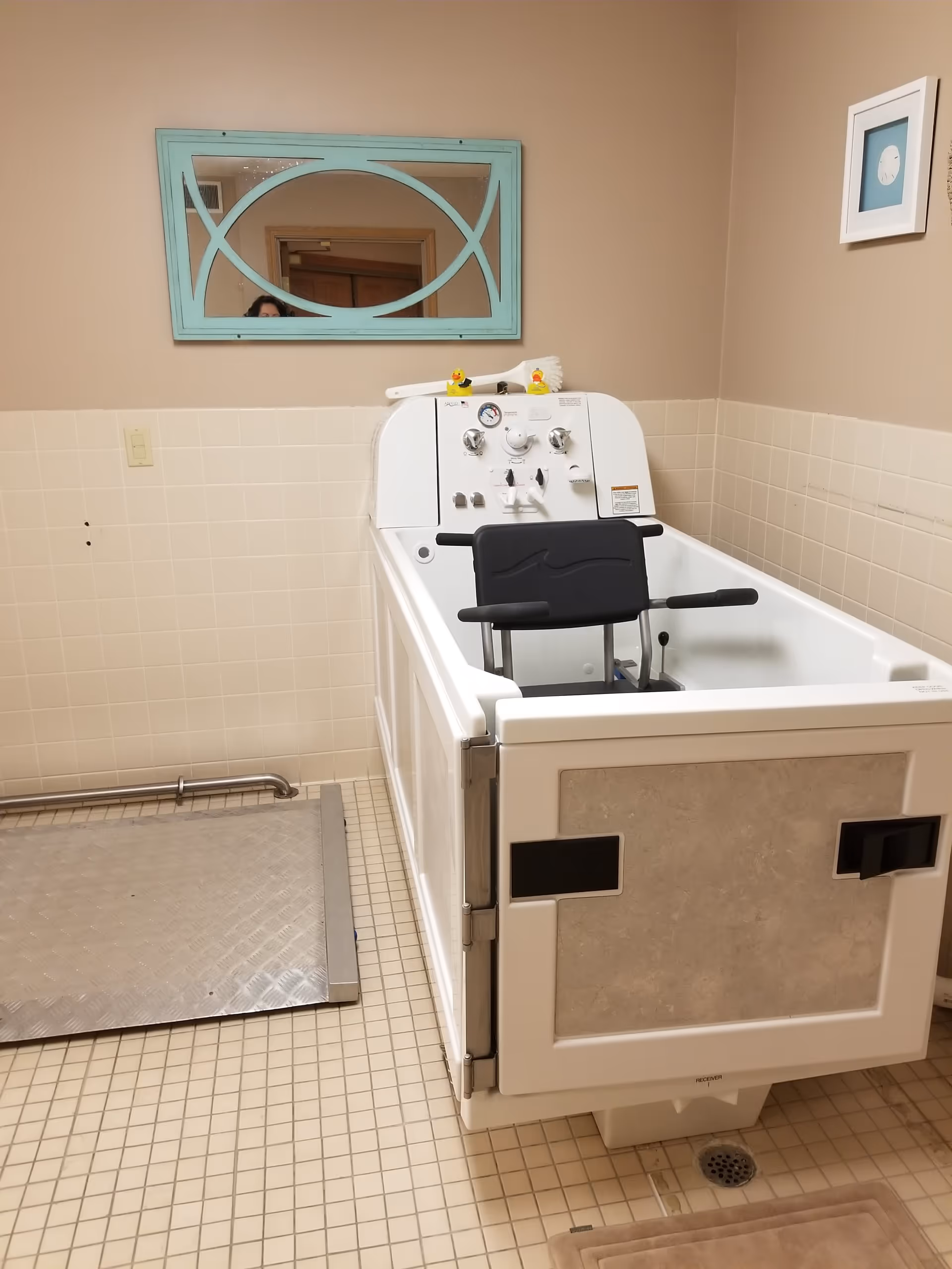 A beige-tiled bathroom with a specialized white bathing tub equipped with a black seat and armrests, control knobs, and two rubber duck toys on top. A turquoise-framed mirror hangs on the wall above the tub, and a small framed clock is mounted on the adjacent wall. There is a metal ramp on the floor next to the tub.