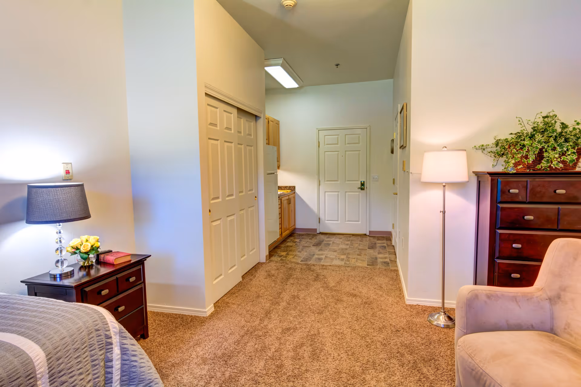 Interior view of a senior living facility room featuring a bed with a bedside table and lamp on the left, a closet with double doors in the center, a small kitchen area with wooden cabinets and a refrigerator, a white entrance door at the back, a floor lamp, a cushioned chair, and a dark wooden dresser with a plant on top on the right.
