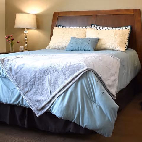 A neatly made bed with a wooden headboard, light blue bedding, a gray blanket folded at the foot, and several pillows including a small blue decorative pillow. To the left of the bed is a nightstand with a lamp and a vase of pink tulips.
