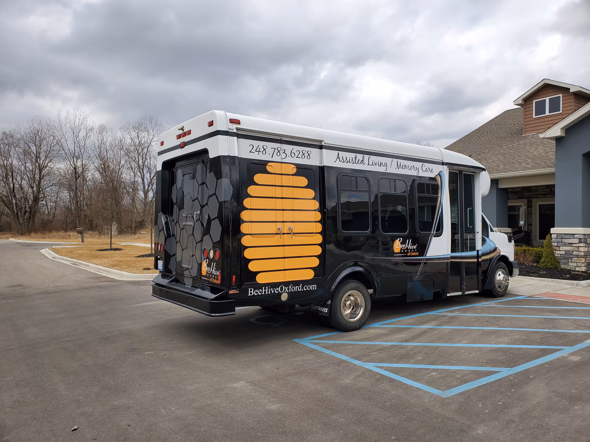 A BeeHive Homes shuttle with a large beehive graphic parked in front of a senior living building under a cloudy sky.