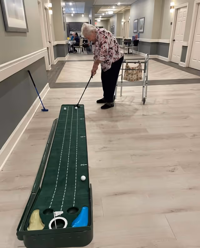 An elderly woman using a walker plays indoor mini golf in a hallway of a senior living facility. The hallway has light-colored wooden floors, gray and white walls, and framed pictures on the walls. In the background, other residents are seated at tables.