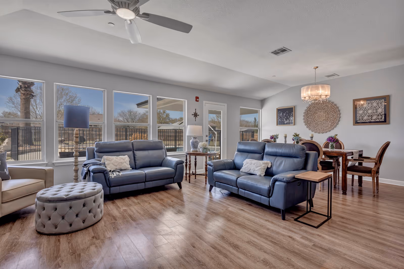 A bright and spacious living room with two blue leather sofas, a beige armchair, and a round tufted ottoman. The room features large windows letting in natural light, a ceiling fan, wooden flooring, and a dining area with a wooden table and chairs in the background. Decorative wall art and a chandelier are visible on the wall behind the dining table.