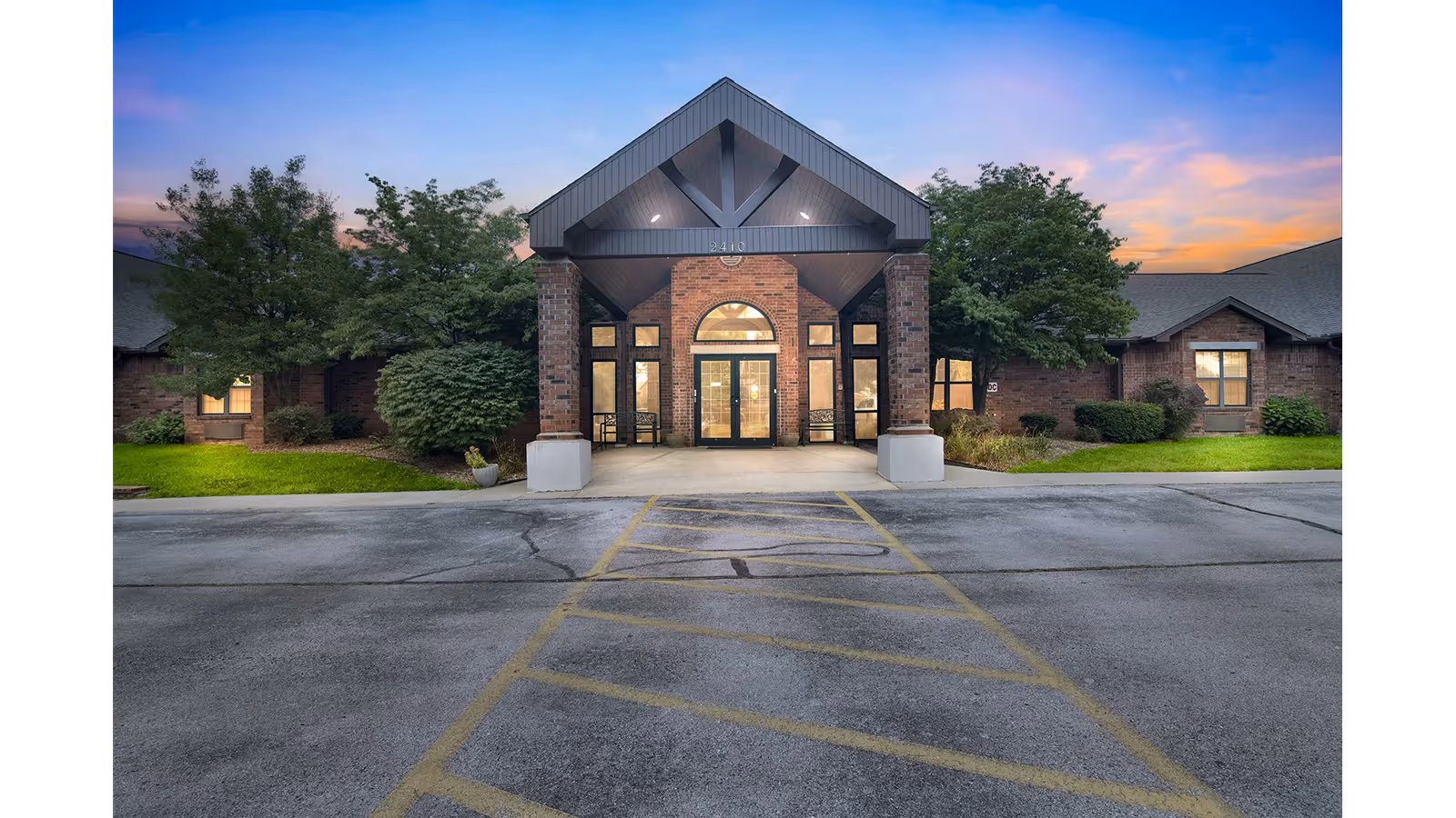 Front exterior view of a brick building with a covered entrance supported by brick columns, surrounded by green trees and shrubs, under a colorful evening sky.