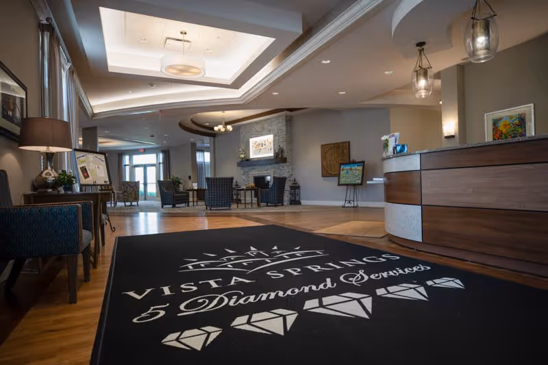 Interior view of a senior living facility lobby with a reception desk on the right, seating area with chairs and tables in the background, a fireplace with artwork above it, and a large black floor mat with the Vista Springs logo and '5 Diamond Services' text.