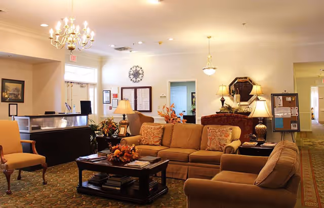 A cozy senior living facility common area with beige sofas, armchairs, a wooden coffee table with magazines and a floral centerpiece, table lamps, a chandelier, a clock on the wall, and a reception desk near the entrance.