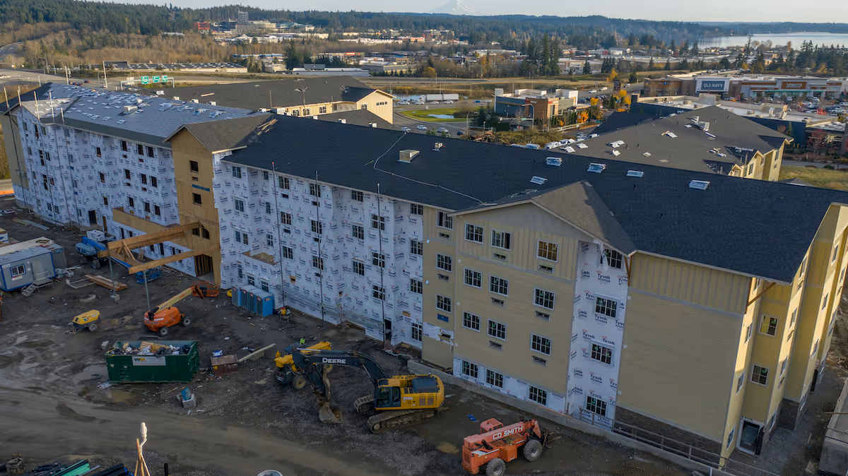 Aerial view of a large multi-story building under construction with construction equipment and materials around the site. The building has a dark roof and partially installed yellow siding, with some areas still covered in white weatherproofing wrap. Surrounding the construction site are roads, commercial buildings, and a distant tree line under a clear sky.
