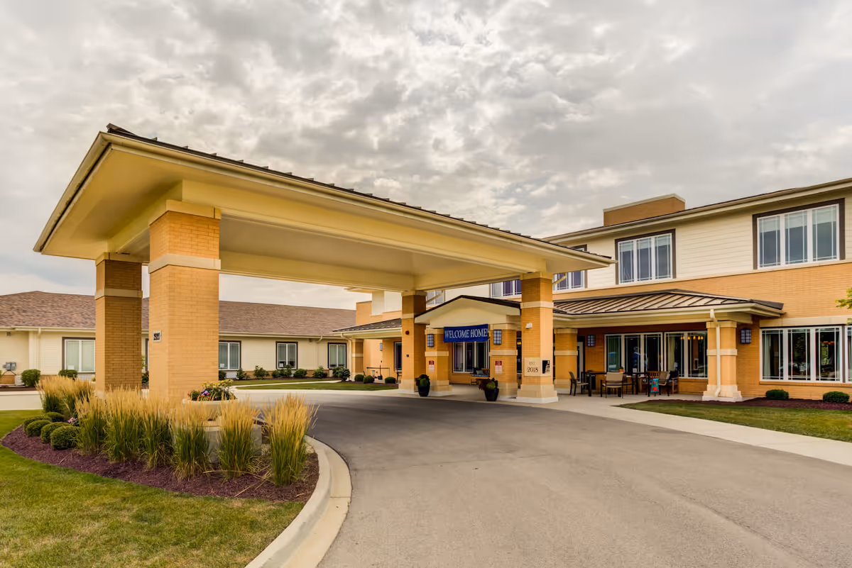 Exterior view of Arbor Terrace Naperville senior living facility showing the entrance with a covered drop-off area, landscaped greenery, and a 'Welcome Home' sign under the canopy.