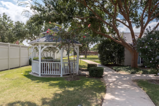 White wooden gazebo next to a paved pathway in a landscaped outdoor courtyard with trees and shrubs.
