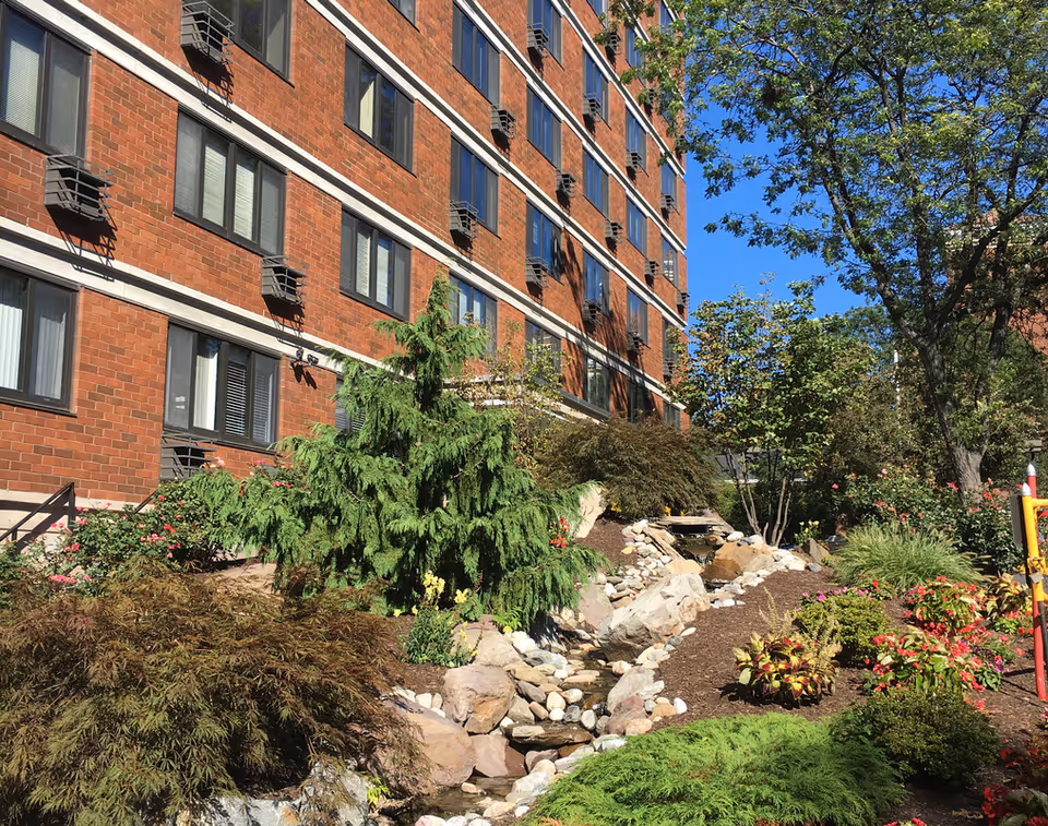 Outdoor garden area next to a multi-story brick building with several windows. The garden features various green shrubs, trees, colorful flowers, rocks, and a small water stream running through the landscaped area under a clear blue sky.