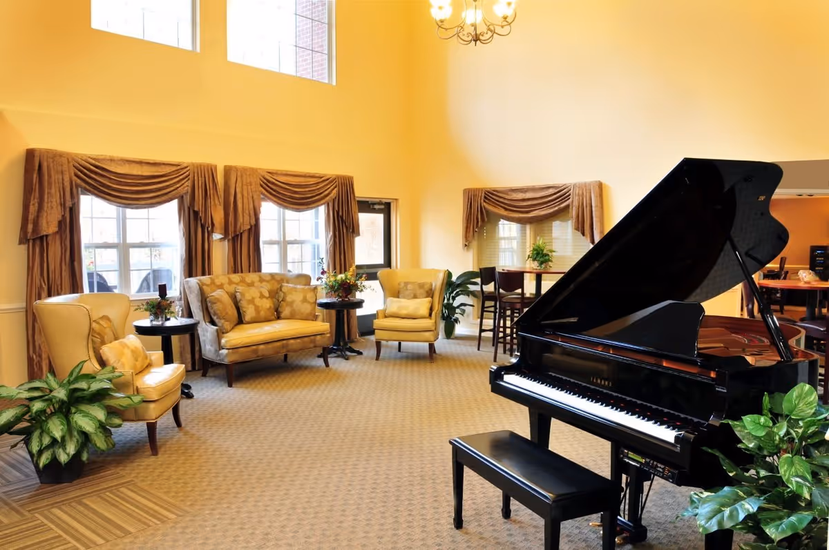 A bright and spacious living room area with high ceilings and large windows covered with brown drapes. The room features a black grand piano with a matching bench, several upholstered chairs and a loveseat with patterned cushions, small round tables with floral arrangements, and green potted plants. A chandelier hangs from the ceiling, and there is a small dining table with chairs in the background.