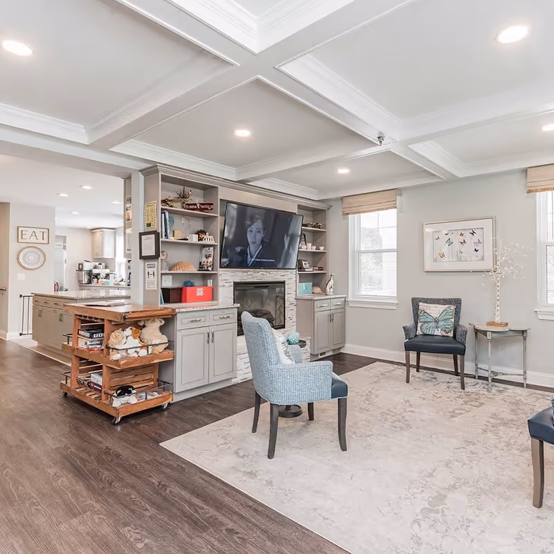 A bright and cozy living room area in a senior living facility with light gray walls, a large area rug, two blue upholstered chairs, a wall-mounted TV above a fireplace, built-in shelves with decorative items, and windows with beige blinds letting in natural light. The room opens into a kitchen area with light cabinetry and a wooden rolling cart with stuffed animals and books.