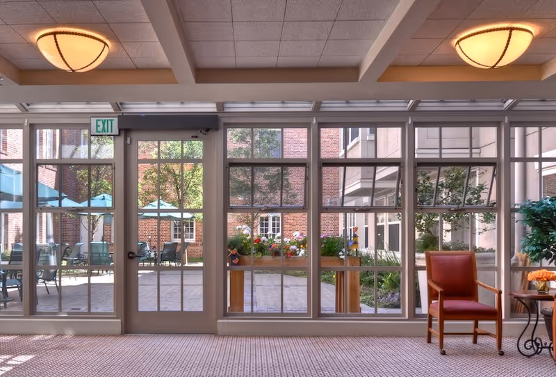 Sunlit interior seating area with large floor-to-ceiling windows looking out onto a brick courtyard with patio tables and umbrellas.