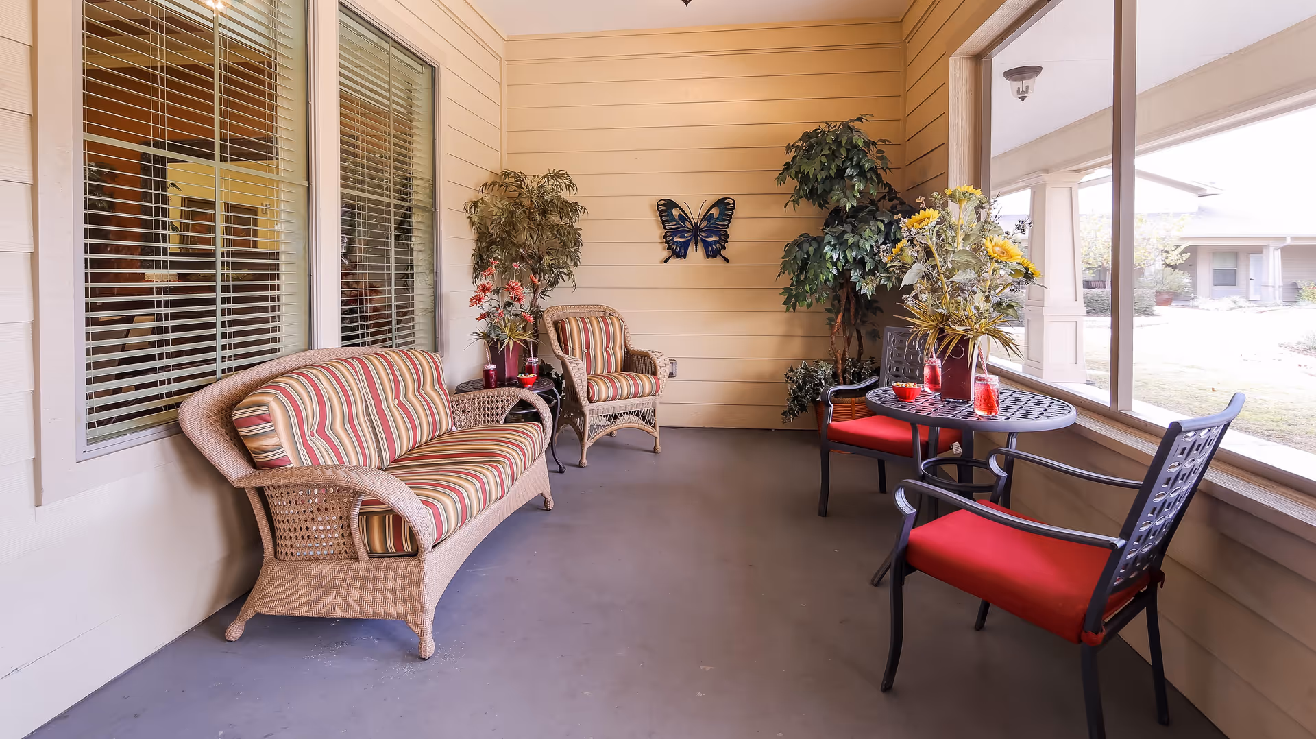 A cozy enclosed porch area with wicker furniture including a striped cushioned loveseat and chair, a small round table with two red cushioned chairs, decorative plants, a butterfly wall decoration, and large windows overlooking an outdoor garden area.