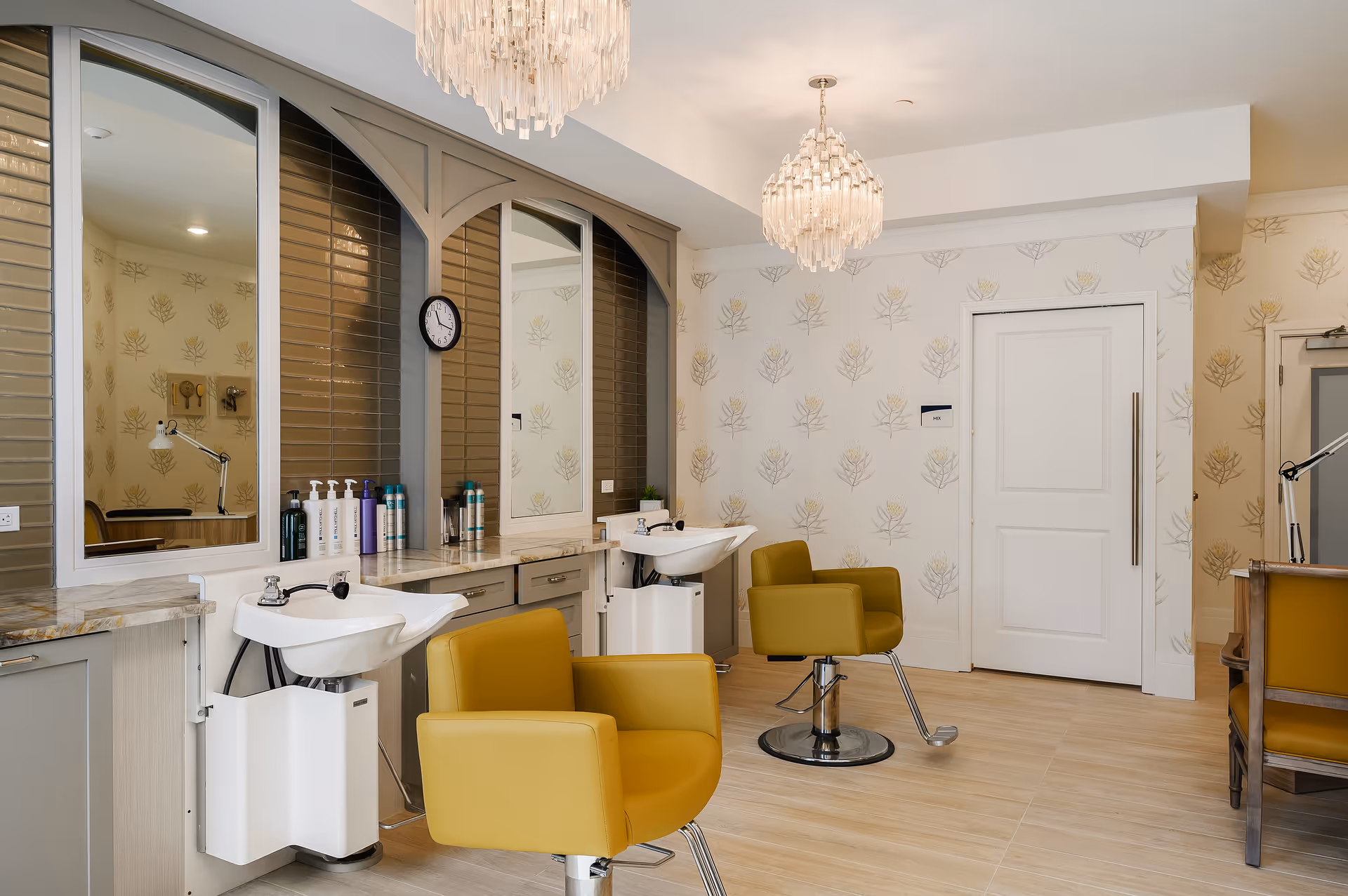 Interior view of a stylish hair salon area in a senior living facility with two yellow salon chairs in front of wash basins, large mirrors, and a countertop with hair care products. The room features light wood flooring, patterned wallpaper, and two elegant chandeliers hanging from the ceiling.