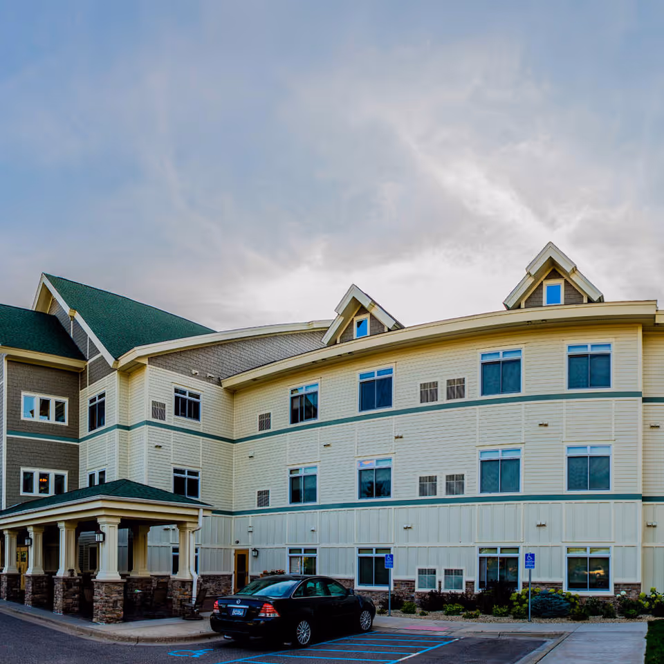 Exterior view of a three-story assisted living facility building with beige and gray siding, green roof shingles, multiple windows, and a covered entrance. A black car is parked in a handicapped parking space in front of the building under a partly cloudy sky.