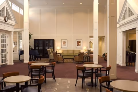 A spacious and well-lit common area with several round tables and chairs in the foreground, and a seating area with a green sofa, two armchairs, a black cabinet, and framed artwork on the wall in the background. The room features high ceilings, large windows, and white pillars.