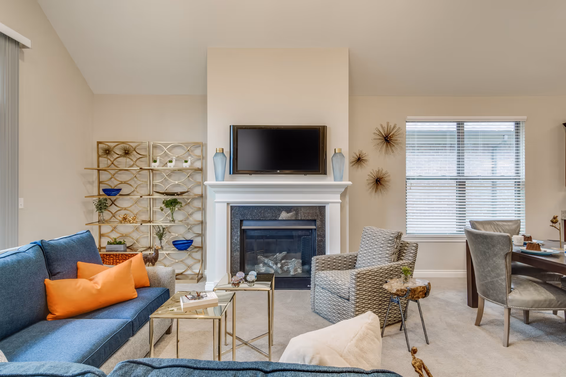 A cozy living room with a blue sofa adorned with orange pillows, a patterned armchair, and a small wooden side table. A white fireplace with a mounted flat-screen TV above it is centered on the wall. Decorative gold wall art and a gold shelving unit with plants and blue bowls are on the left side. A window with blinds and a dining table with chairs are visible on the right side.